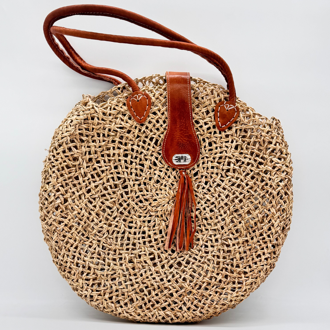Runde, handgewebte Basttasche aus grobem Raffia mit braunem Lederüberschlag, Quastenanhänger und zwei Lederhenkeln