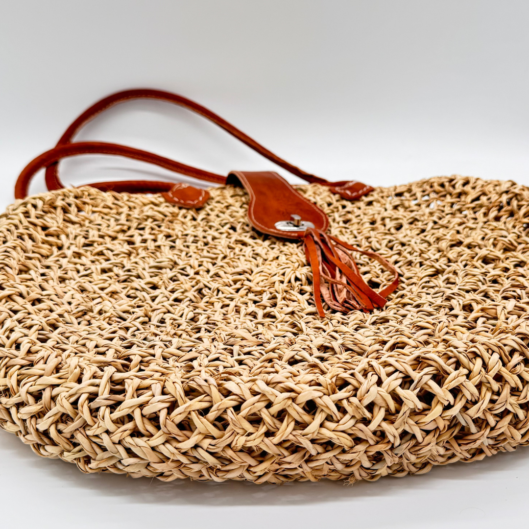 Runde, handgewebte Basttasche aus grobem Raffia mit braunem Lederüberschlag, Quastenanhänger und zwei Lederhenkeln
