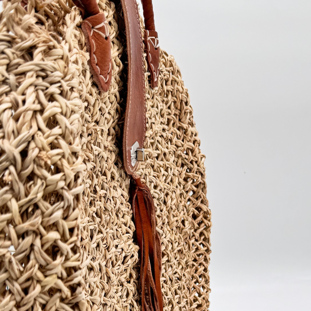 Runde, handgewebte Basttasche aus grobem Raffia mit braunem Lederüberschlag, Quastenanhänger und zwei Lederhenkeln