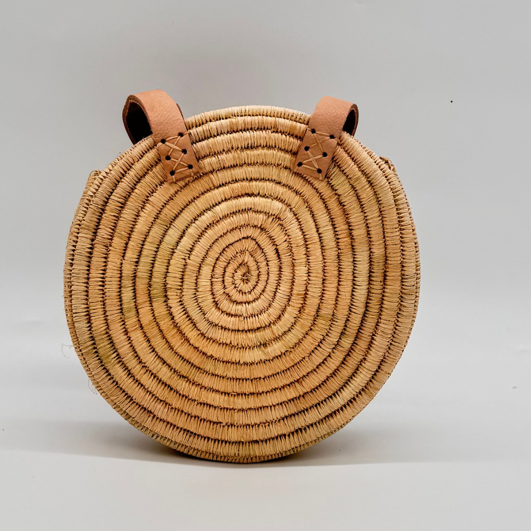 Kleine, runde Basttasche aus handgewebtem Raffia mit zwei braunen Lederlaschen zur Befestigung eines Trageriemens