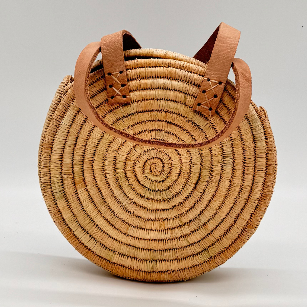 Kleine, runde Basttasche aus handgewebtem Raffia mit zwei braunen Lederlaschen zur Befestigung eines Trageriemens