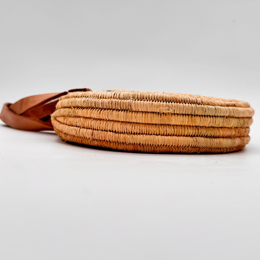 Kleine, runde Basttasche aus handgewebtem Raffia mit zwei braunen Lederlaschen zur Befestigung eines Trageriemens