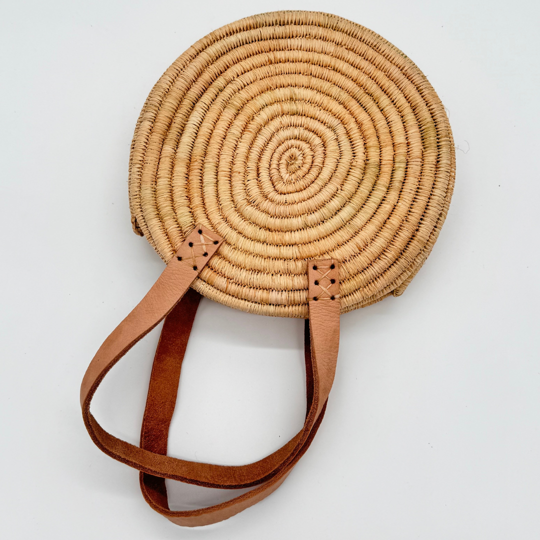 Kleine, runde Basttasche aus handgewebtem Raffia mit zwei braunen Lederlaschen zur Befestigung eines Trageriemens