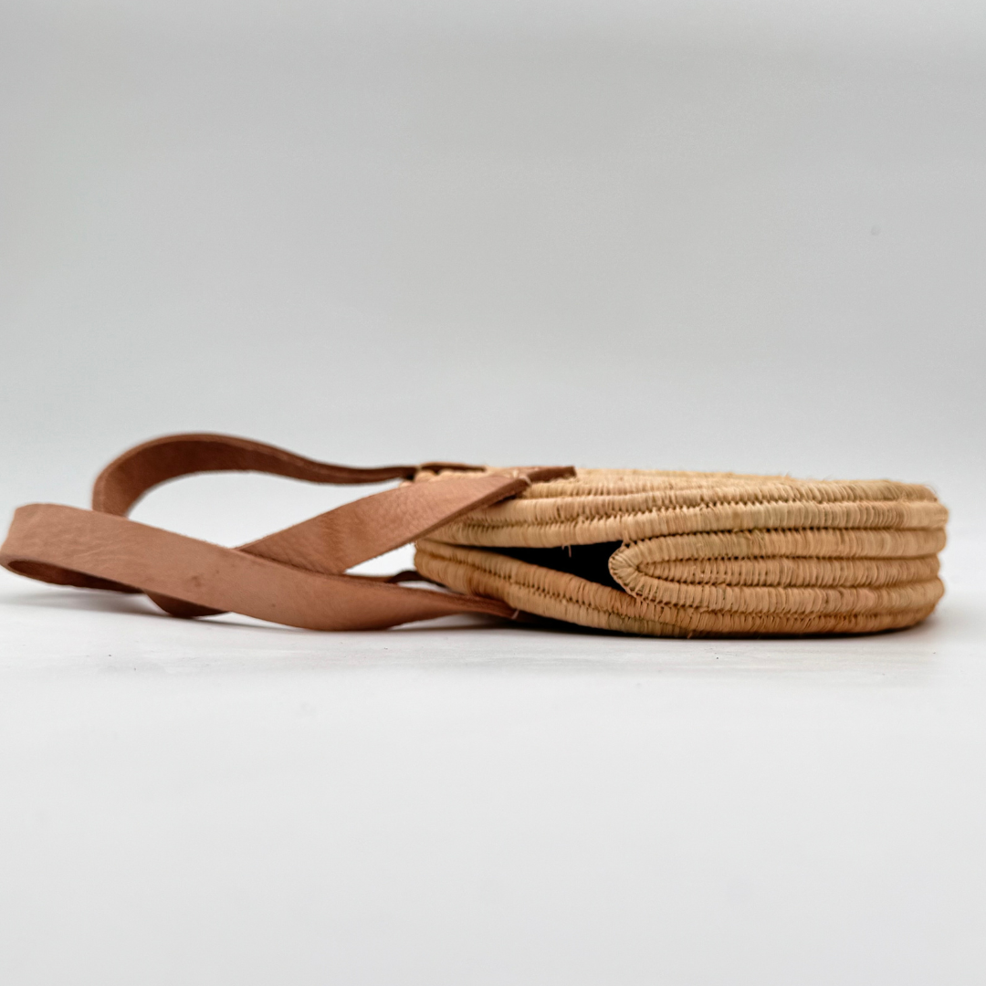 Kleine, runde Basttasche aus handgewebtem Raffia mit zwei braunen Lederlaschen zur Befestigung eines Trageriemens