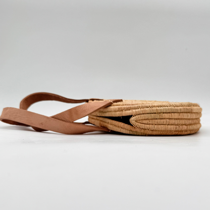 Kleine, runde Basttasche aus handgewebtem Raffia mit zwei braunen Lederlaschen zur Befestigung eines Trageriemens