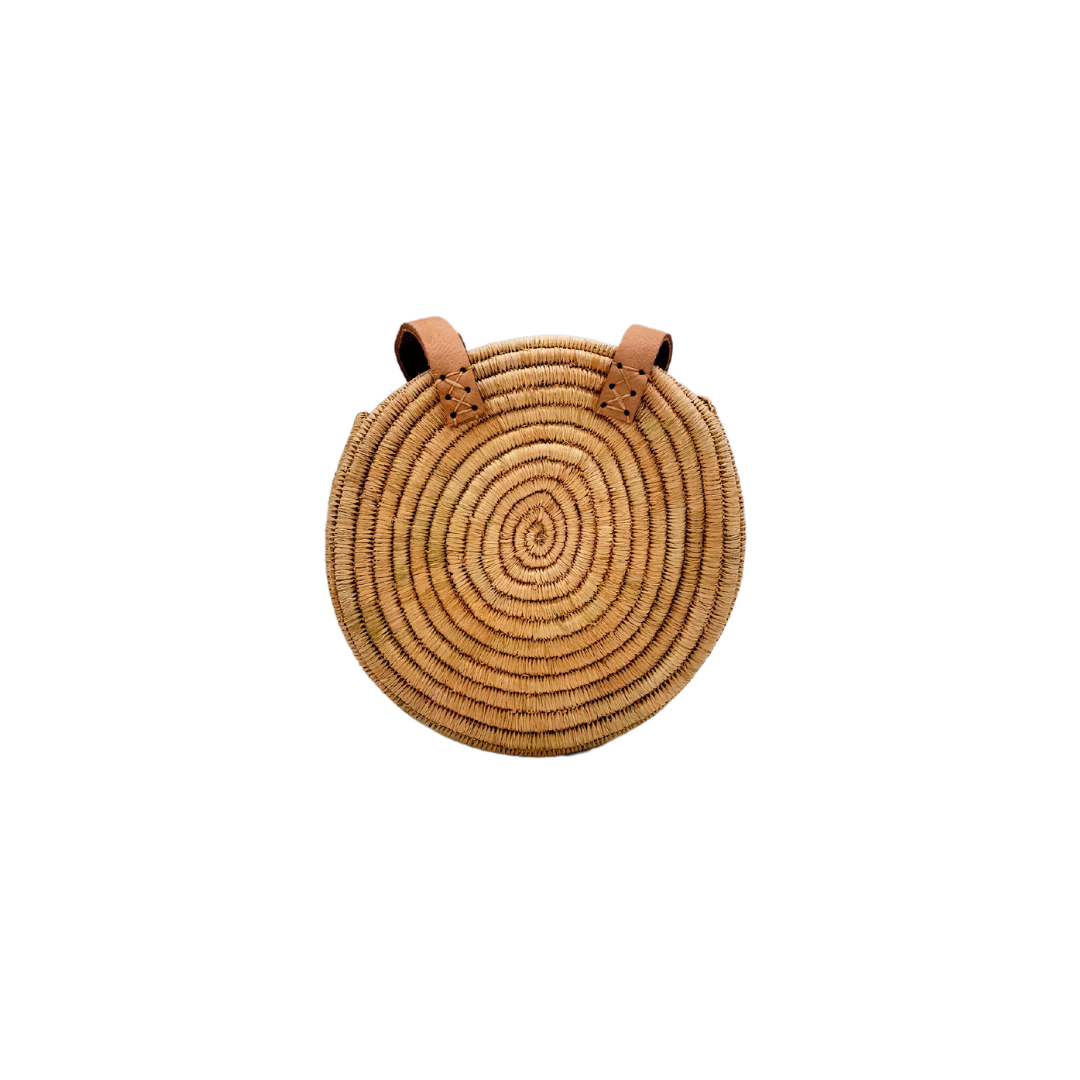 Kleine, runde Basttasche aus handgewebtem Raffia mit zwei braunen Lederlaschen zur Befestigung eines Trageriemens