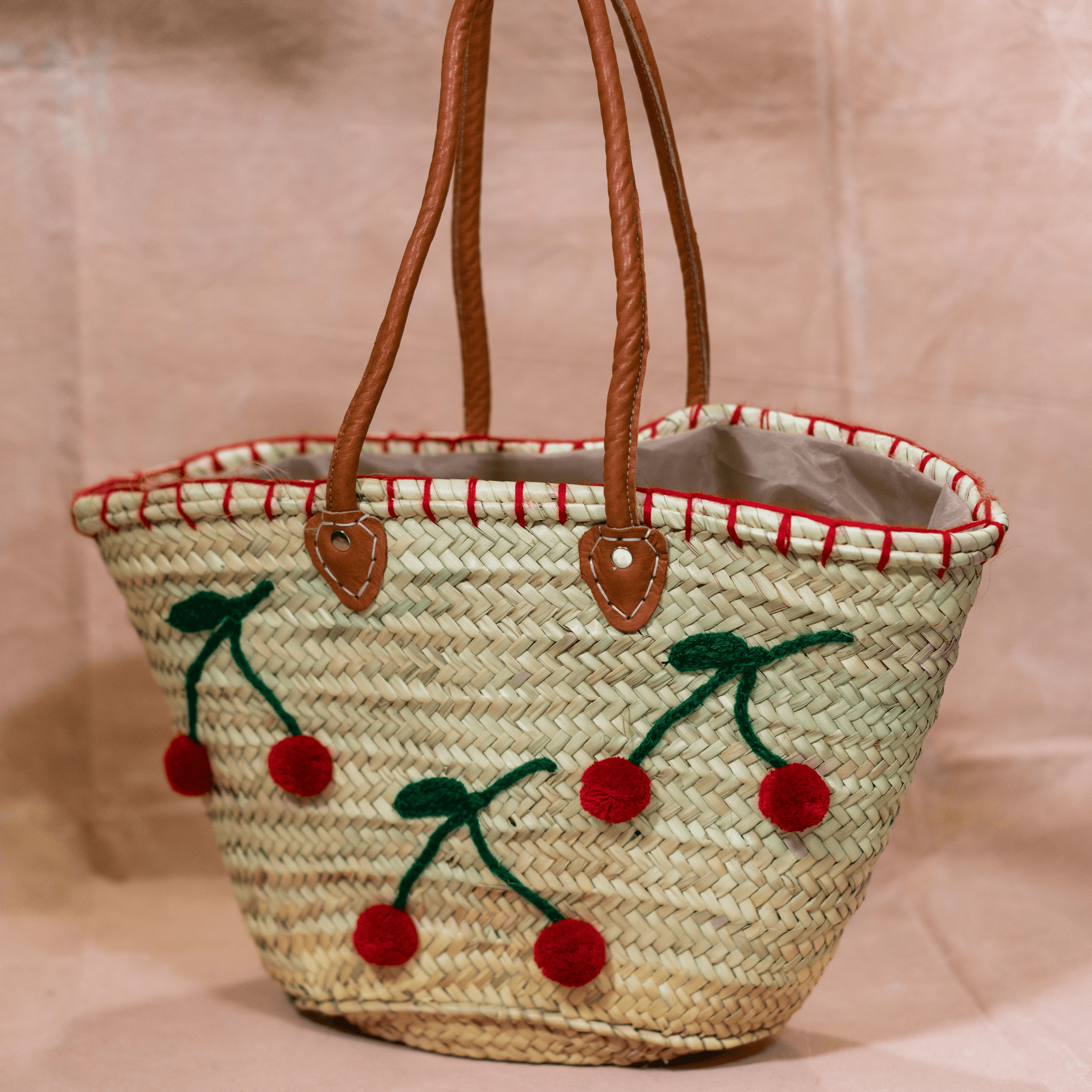 Handgewebte Basttasche aus Naturstroh mit grünen Garnstielen und roten Pompon-Kirschen sowie braunen Lederhenkeln