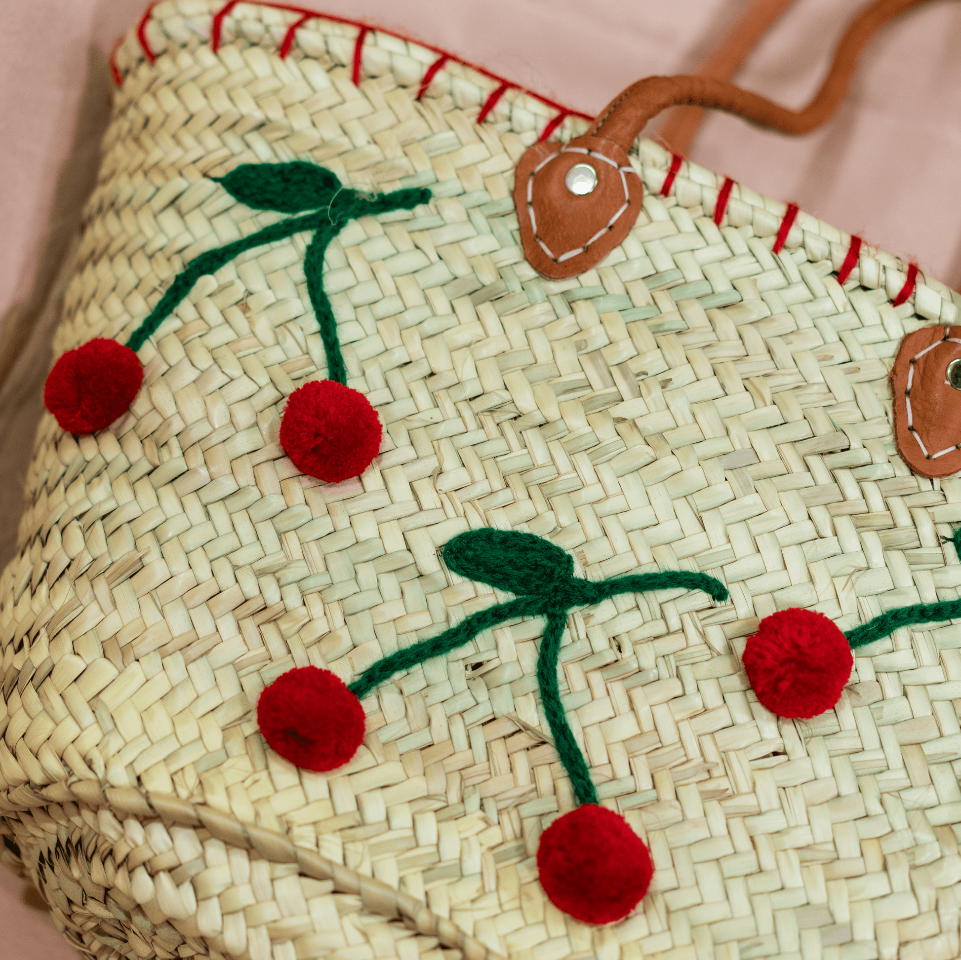 Handgewebte Basttasche aus Naturstroh mit grünen Garnstielen und roten Pompon-Kirschen sowie braunen Lederhenkeln