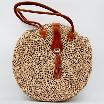 Runde, handgewebte Basttasche aus grobem Raffia mit braunem Lederüberschlag, Quastenanhänger und zwei Lederhenkeln